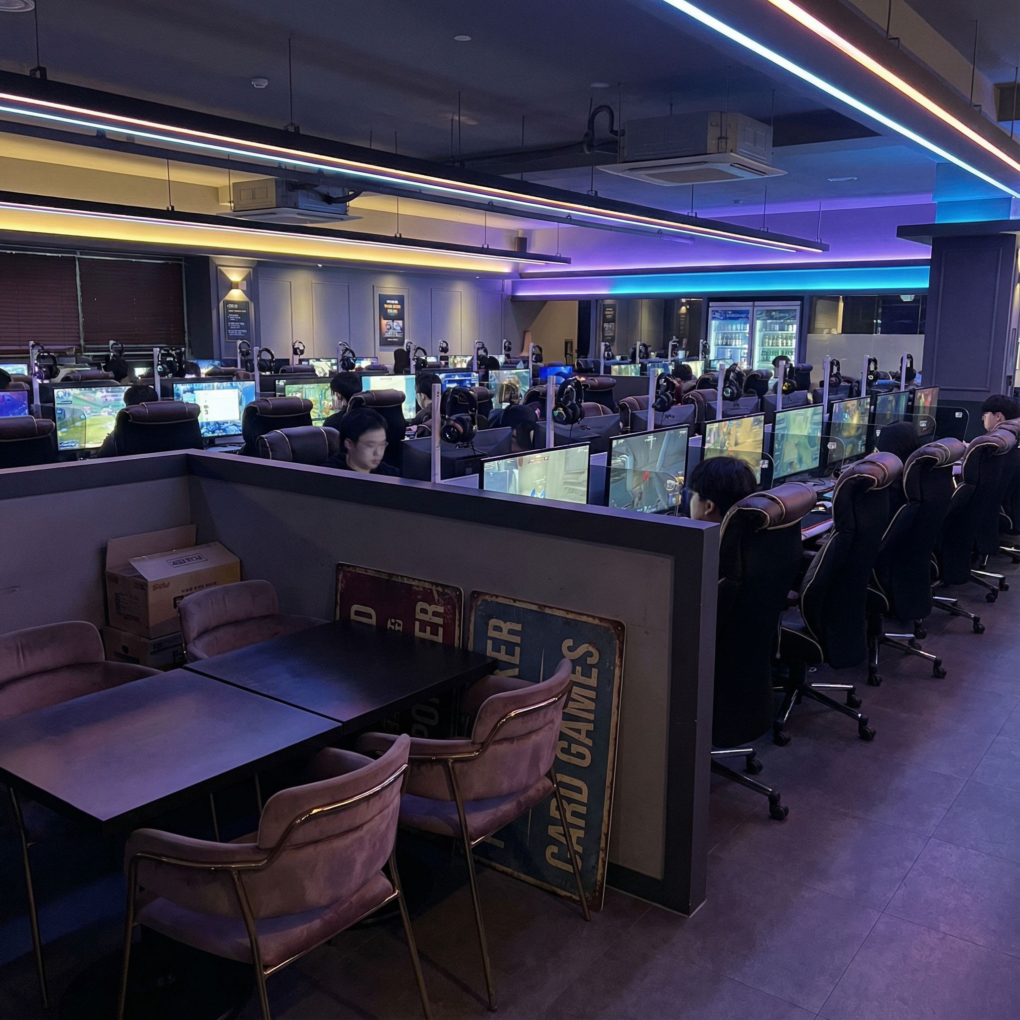 어두운 조명 속 PC방, 게임용 PC 줄지어 있고 카드게임 표지 뗀 빈 예약석 모습이다